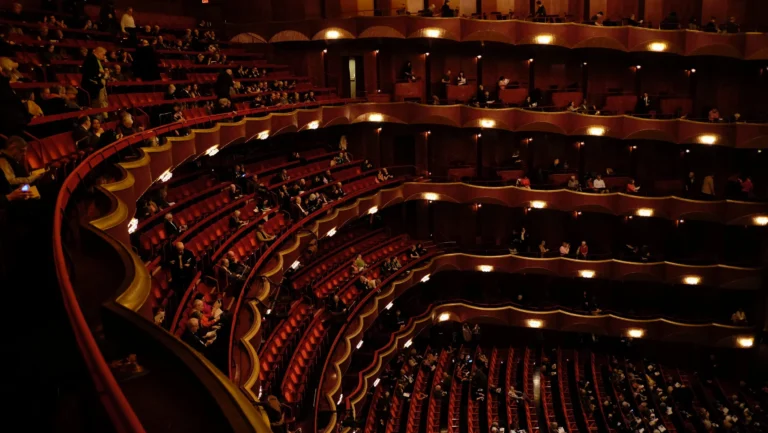 A raised view of an upmarket theatre venue, rings of red seats on multiple levels of balconies looking down towards the stage.