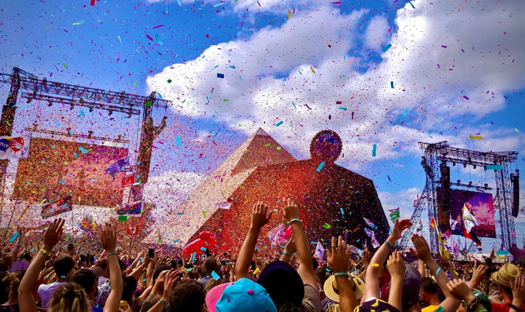 A wide shot of a crowd at a festival, everyone with their arms waving in the air. Behind them is a festival stage, set against a blue sky with some clouds drifting overhead. The air is full of confetti.