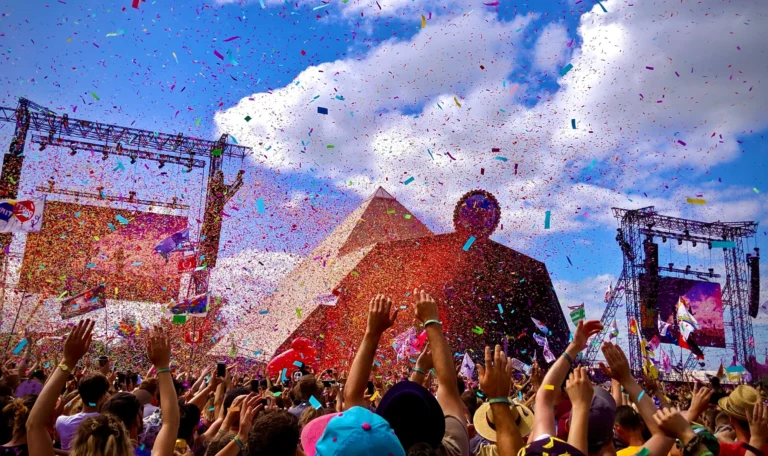 A wide shot of a crowd at a festival, everyone with their arms waving in the air. Behind them is a festival stage, set against a blue sky with some clouds drifting overhead. The air is full of confetti.