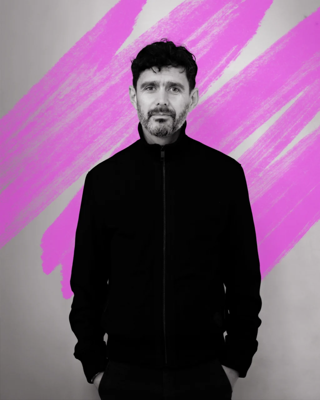 a black and white portrait of a man wearing a black jumper, zipped up to his chin. Behind him is a hand-drawn scribble in neon pink.