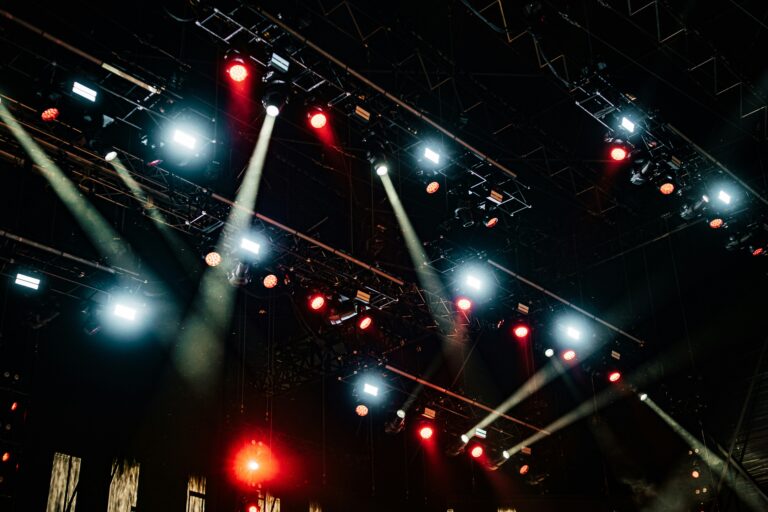 a dark image of stage lights and scaffolding above a stage, seen from below. the lights are red and blue and white, all pointing in different directions.