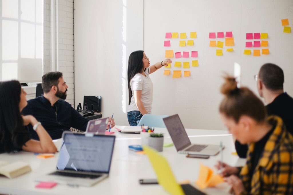 A group of people sat in a meeting room. A woman is putting multi-coloured sticky notes up on the wall, while the rest of the people are at a desk writing on sticky notes or looking at their laptops.