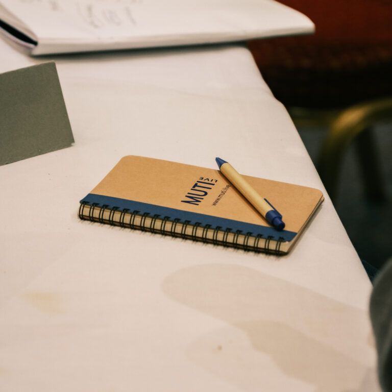 A brown and blue MUTI Live branded notebook laying closed on a white desk. Other pieces of paper and notebooks are cut off at the edge of the image.