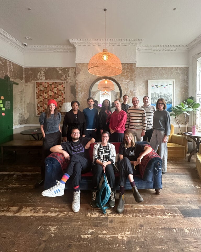 A diverse group of people sat on, and stood behind, a blue sofa in a vintage-styled hotel room, all looking at the camera.