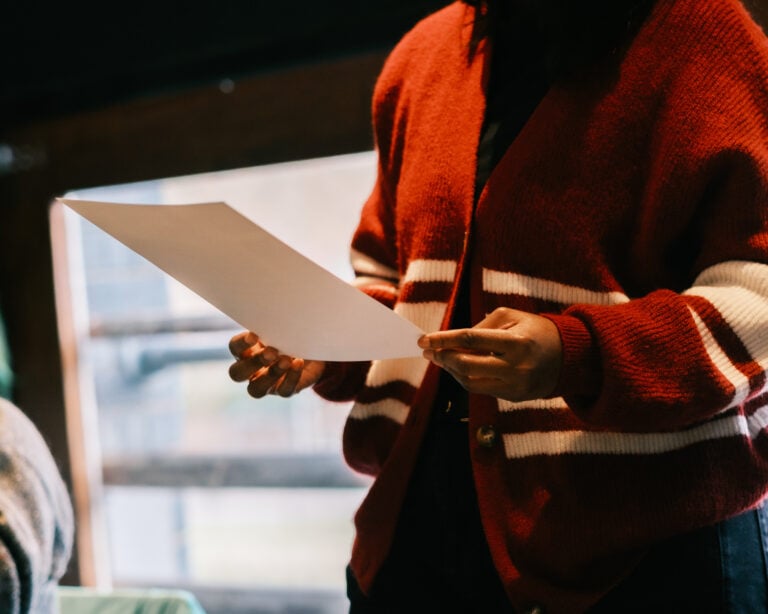 A woman's hands holding piece of paper, studying it. She has dark skin, and is wearing a red and white cardigan. She's standing in front of a window.