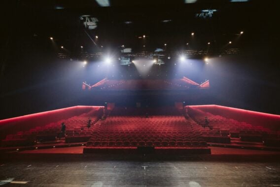 An empty theatre auditorium from the point of view of the stage, looking out at rows of empty red seats bathed in cold light from spotlights hanging from the ceiling.