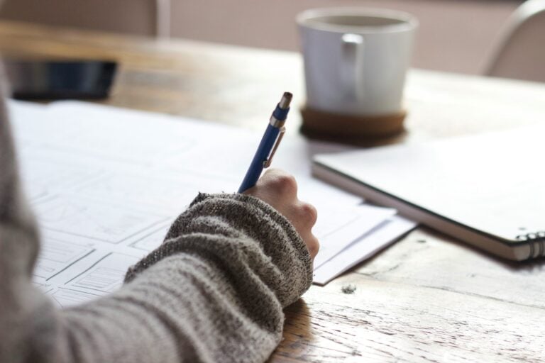 A close up a person's hand making notes on a large notepad. They're wearing a cardigan. Out of focus on the table is a mug of coffee and another notebook.