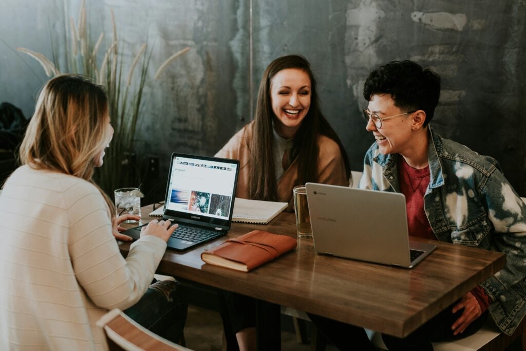 three diverse women sat around a desk, laughing while typing and looking at their laptops.