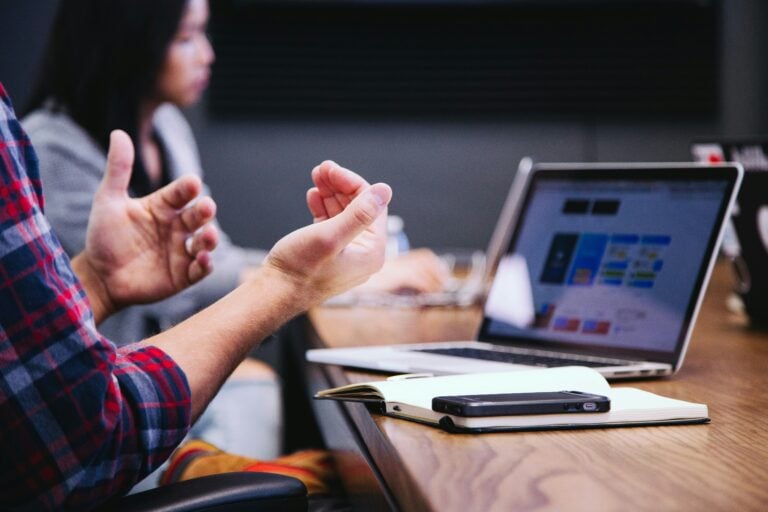 A close up of hands gesturing as if someone is speaking. On the desk in front of them is an open notebook and phone, face down. The background is out of focus, but there is someone else sat in the back of the image.